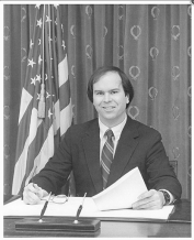 A person sitting at a desk with a flag behind him

Description automatically generated