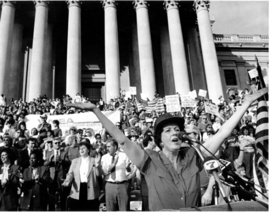A person speaking into a microphone with her arms raised

Description automatically generated
